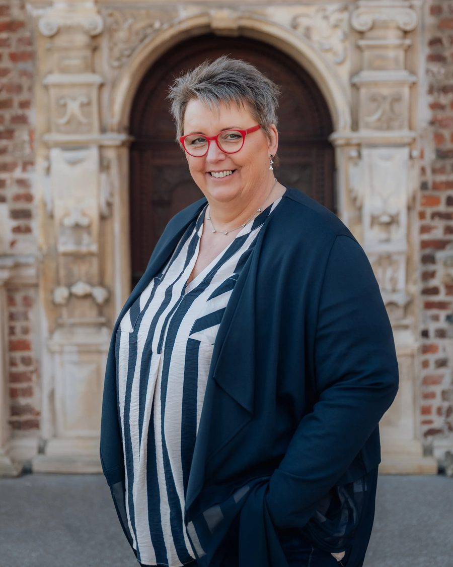 Christel Schilderink, kurze graue Haare und rote Brille, steht lächelnd vor einer alten Bogentür